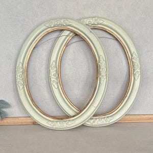 May include: Two oval picture frames with a pale sage green finish and gold-toned inner rims. The frames feature raised floral carvings at the top, bottom, and sides. Each frame includes a thin wire attached to the back for hanging purposes.