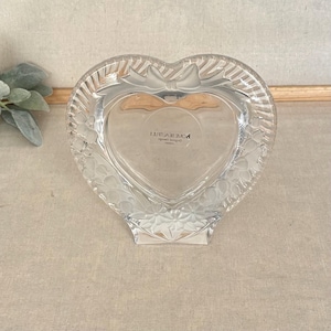 May include: A heart-shaped clear glass picture frame featuring a decorative bow at the top and embossed floral patterns along the edges. The center of the frame includes a sticker with the text HOME BEAUTIFUL Original Design Japan.