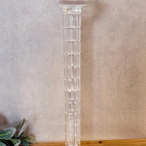 May include: A clear, glass pedestal with a square base and top. The column is rectangular with a repeating pattern. The pedestal is approximately 12 inches tall, suitable for displaying decorative items.