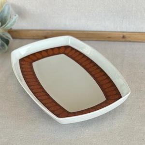 May include: A white rectangular serving dish with a brown leaf pattern border. The dish has a slightly curved shape and is set against a neutral background. The interior of the dish is white.