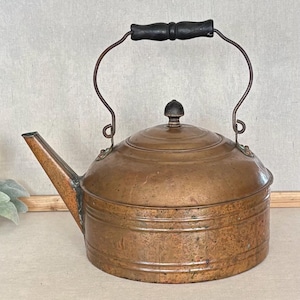 May include: A vintage copper tea kettle with a cylindrical body, a wide spout, and a rounded lid featuring a small knob. The kettle has a thin metal handle arched over the top, equipped with a black wooden grip for handling the vessel.