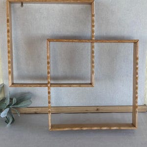 May include: Two overlapping square wooden wall shelves with a light brown finish and a scalloped, wavy edge detail along the outer frame. The shelves are constructed from smooth wood and feature a simple, open-box design for wall mounting.