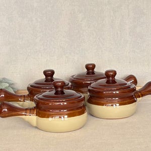 May include: A set of three ceramic soup crocks with lids, each featuring a two-tone design with a dark brown glazed upper section and a light beige lower section. Each crock includes a single side handle and a matching removable lid with a knob.