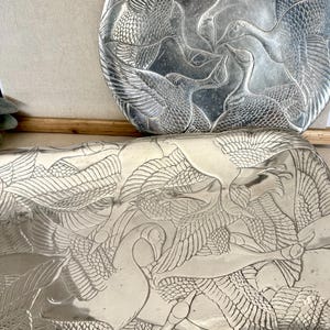 May include: A silver-toned metal tray and a matching circular plate, both featuring an intricate embossed pattern of multiple geese in flight with detailed wing feathers and long necks, creating a cohesive metallic set with a reflective finish.