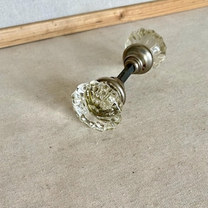 May include: A clear glass door knob set featuring a faceted, multi-sided design on both ends. The two glass knobs are connected by a central metal spindle with a brushed nickel finish, creating a functional hardware piece for doors.