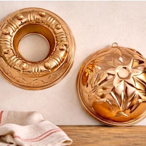 May include: Two copper-colored bundt cake pans. One is round with a central hole and decorative ridges. The other is a dome shape with a starburst design. A white and red striped cloth is in the foreground.