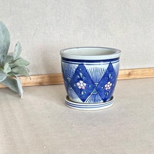 May include: A ceramic plant pot with a matching saucer featuring a blue and white geometric diamond pattern. The design includes radiating lines and small pink and green floral accents centered within each diamond shape on the exterior surface.