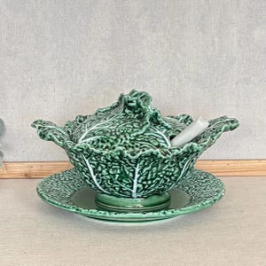 May include: A green ceramic tureen shaped like a cabbage head with a matching lid and saucer. The tureen features textured, ruffled leaf edges and a white ceramic ladle resting inside the bowl, with a glossy finish throughout the entire set.