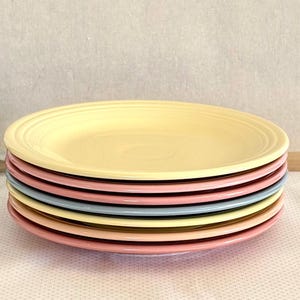 May include: A stack of seven colorful ceramic plates. The top plate is a pale yellow, with the other plates in shades of pink, blue, and yellow. The plates have a simple, classic design.