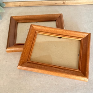 May include: Two rectangular picture frames made of medium-toned wood with a smooth, polished finish. Each frame features a wide, beveled border and contains a clear glass pane protecting a light tan backing board with a visible easel stand.
