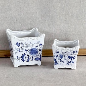May include: Two white ceramic planters with a blue floral design. The planters are square-shaped with a scalloped rim and a blue floral pattern. The larger planter is approximately 6 inches tall, and the smaller one is about 4 inches tall.