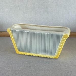 May include: A rectangular ceramic planter featuring a central panel with vertical gray and white stripes. The planter is framed by a bright yellow, rope-textured border along the top and bottom edges, with a smooth, off-white interior basin.
