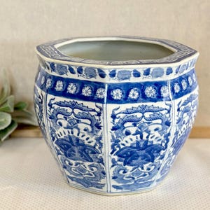 May include: A blue and white ceramic planter with an octagonal shape. The planter features intricate floral and foliate patterns in blue against a white background. The rim has a decorative border, and the interior is a light gray color. A decorative piece for home decor.