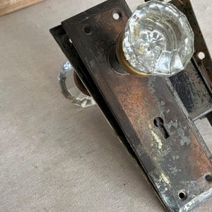 May include: A vintage door handle set featuring two clear, faceted glass knobs attached to a rectangular, weathered metal backplate with a central keyhole. The metal plate shows signs of oxidation and wear, with visible screw holes at the top and bottom.