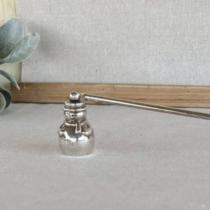 May include: A silver-toned metal candle wick trimmer featuring a handle and a bell-shaped snuffer end. The snuffer is shaped like a small snowman wearing a hat and a scarf, with a smooth, reflective metallic finish throughout the entire object.