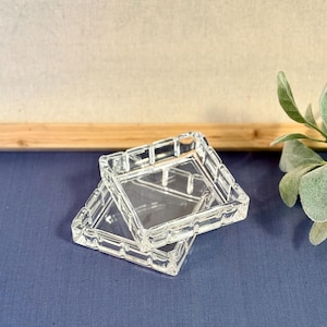 Puede incluir: Dos ceniceros cuadrados de vidrio transparente apilados sobre una superficie azul. Los ceniceros tienen un diseño de rejilla en los bordes y un centro claro y abierto. El fondo es un lienzo neutro con un marco de madera.