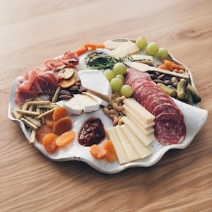 Puede incluir: Una fuente blanca con bordes ondulados llena de una variedad de aperitivos. La fuente incluye salami en rodajas, queso, uvas, nueces, albaricoques secos, galletas saladas y pepinillos. La comida está dispuesta sobre una superficie de madera clara.