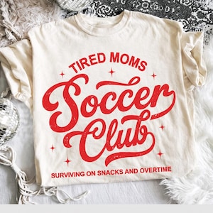 May include: Cream-colored t-shirt with the words "TIRED MOMS Soccer Club" in red, stylized lettering. The text "SURVIVING ON SNACKS AND OVERTIME" is printed below. The shirt has short sleeves and a relaxed fit.