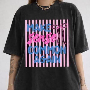 May include: Black t-shirt with the text "MAKE sense COMMON AGAIN" in blue and pink, set against a pink and white striped background. A pink bow is on the right side of the text. The shirt has short sleeves and a crew neck.