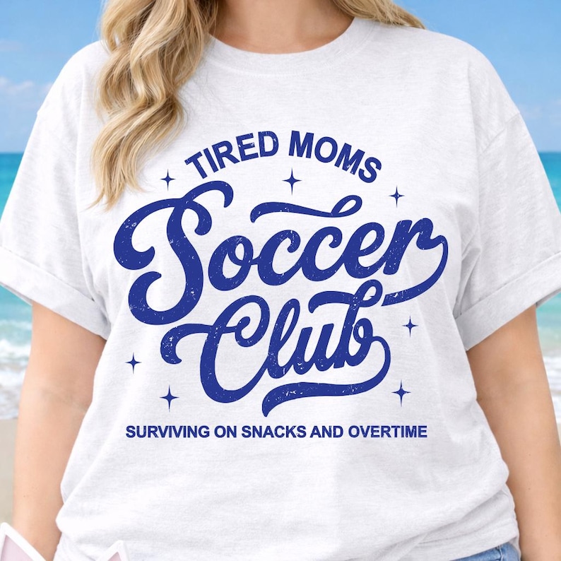 May include: White t-shirt with the text "TIRED MOMS Soccer Club" in blue, with the phrase "SURVIVING ON SNACKS AND OVERTIME" below. The design includes star accents. The shirt has rolled-up sleeves.