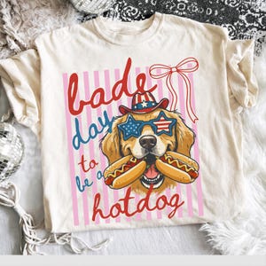 May include: Cream-coloured t-shirt featuring a golden retriever wearing patriotic sunglasses and a hat, holding hot dogs. The text reads "Bad day to be a hotdog" in red and blue.