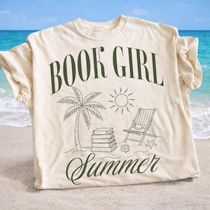 May include: Cream-colored t-shirt with the words "BOOK GIRL Summer" in dark green script. The design includes a palm tree, sun, beach chair, books, and seashells, evoking a summer beach reading theme.