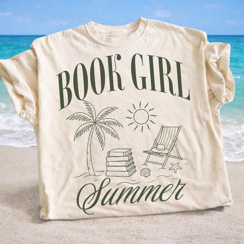 May include: Cream-colored t-shirt with the words "BOOK GIRL Summer" in dark green script. The design includes a palm tree, sun, beach chair, books, and seashells, evoking a summer beach reading theme.