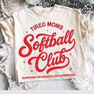 Può includere: T-shirt color crema con la scritta rossa "TIRED MOMS Softball Club" in un carattere invecchiato. La frase "SURVIVING ON SNACKS AND HOMERUNS" è stampata sotto il nome del club. La maglietta ha maniche corte.