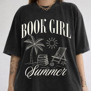 May include: Black t-shirt with the words "BOOK GIRL Summer" in a white, stylized font. The graphic features a palm tree, sun, books, beach chair, and seashell, evoking a summer reading theme.