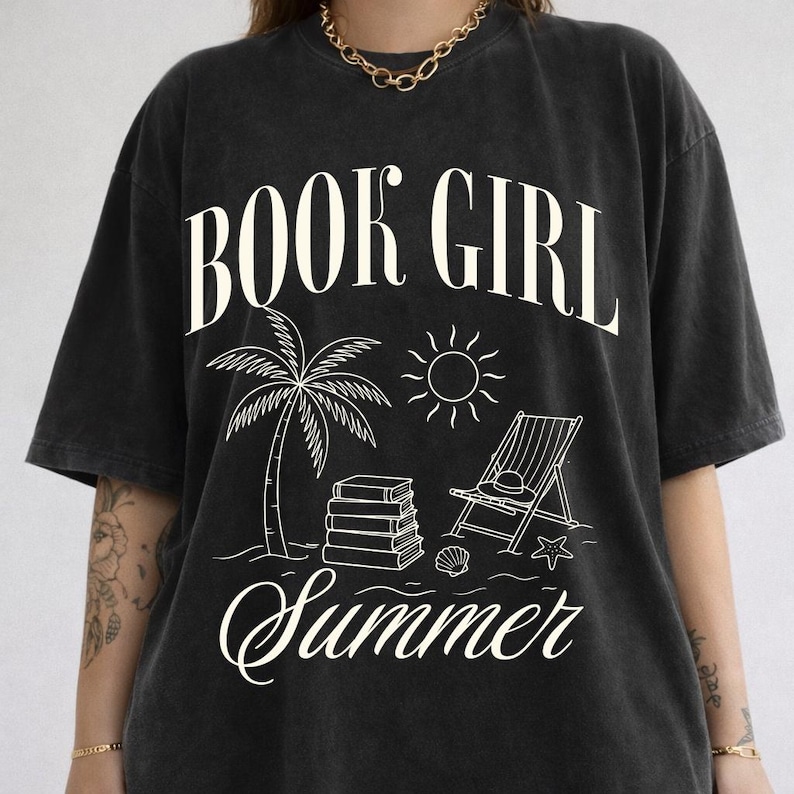 May include: Black t-shirt with the words "BOOK GIRL Summer" in a white, stylized font. The graphic features a palm tree, sun, books, beach chair, and seashell, evoking a summer reading theme.