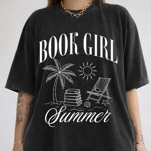 May include: Black t-shirt with a white graphic that reads "BOOK GIRL Summer." The design includes a palm tree, sun, beach chair, books, and seashells. The shirt has a relaxed fit and short sleeves.