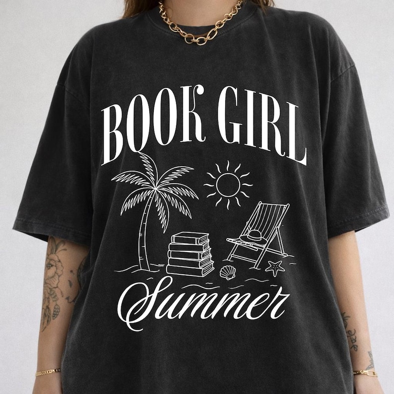 May include: Black t-shirt with a white graphic that reads "BOOK GIRL Summer." The design includes a palm tree, sun, beach chair, books, and seashells. The shirt has a relaxed fit and short sleeves.