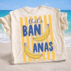 May include: Cream-colored t-shirt with the phrase "that's BANANAS" in blue, featuring two yellow bananas. The design includes vertical yellow stripes. The shirt is displayed on a sandy beach with the ocean in the background.