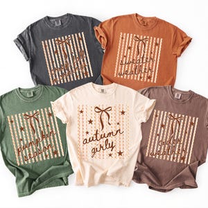 May include: Five short-sleeve crew neck t-shirts in charcoal, rust, olive green, cream, and brown. Each shirt features a central graphic of a bow, stars, and vertical stripes with text reading crisp fall mornings, sweater weather, pumpkin season, autumn girly, and cider weather.