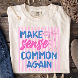 May include: Cream-colored t-shirt with the phrase "MAKE sense COMMON AGAIN" in blue and pink, set against a pink striped background. A pink bow graphic is also present. The shirt is folded on a wooden surface.
