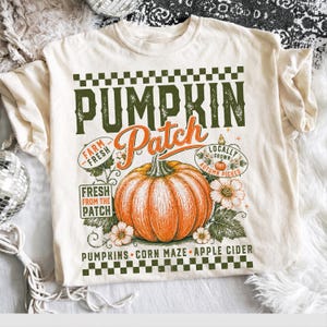 Pode incluir: Uma t-shirt de cor creme com uma impressão gráfica que mostra uma abóbora laranja grande, flores brancas e folhas verdes. O texto diz PUMPKIN PATCH, FARM FRESH, LOCALLY GROWN, AUTUMN PICKED, FRESH FROM THE PATCH, PUMPKINS, CORN MAZE, APPLE CIDER.