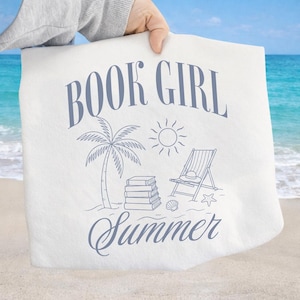 May include: White tote bag with a blue illustration featuring the words "BOOK GIRL Summer". The design includes a palm tree, sun, beach chair, books, seashell, and starfish. The bag is held up against a beach background.