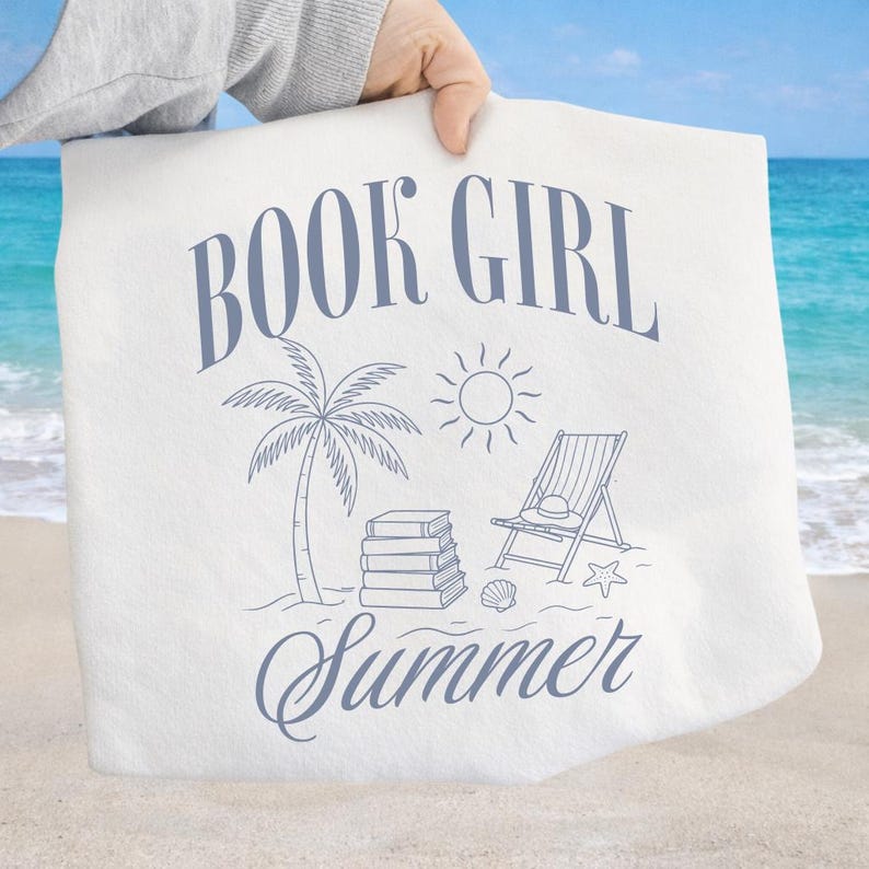 May include: White tote bag with a blue illustration featuring the words "BOOK GIRL Summer". The design includes a palm tree, sun, beach chair, books, seashell, and starfish. The bag is held up against a beach background.