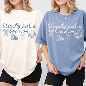Puede incluir: Dos camisetas, una crema y otra azul, con el texto "literally just a boy mom". El diseño incluye un lazo, una excavadora, otro lazo y balones deportivos. Las camisetas se combinan con pantalones cortos negros.