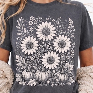 May include: A charcoal gray short-sleeve t-shirt featuring a cream-colored graphic print of three large sunflowers, smaller wildflowers, three pumpkins, and two acorns arranged in a botanical composition on the front of the garment.