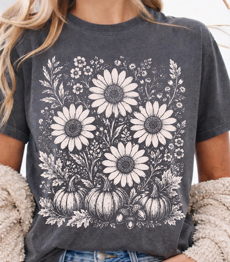May include: A charcoal gray short-sleeve t-shirt featuring a cream-colored graphic print of three large sunflowers, smaller wildflowers, three pumpkins, and two acorns arranged in a botanical composition on the front of the garment.