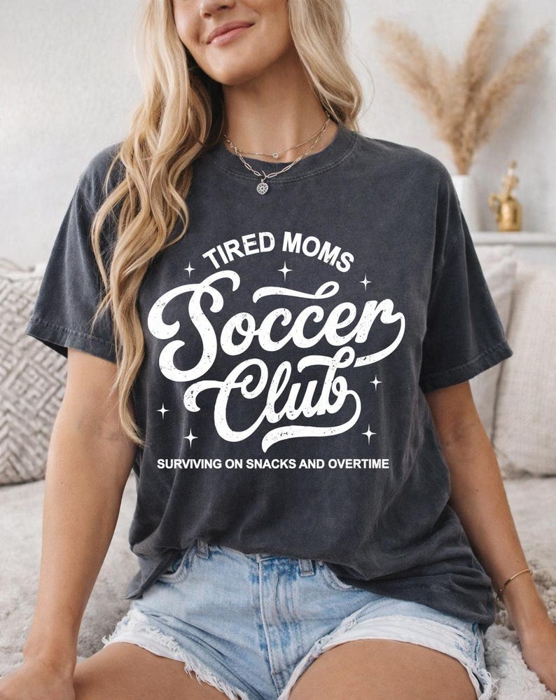 May include: Dark gray t-shirt with the text "TIRED MOMS Soccer Club" in white script. The shirt also includes the phrase "SURVIVING ON SNACKS AND OVERTIME" in a smaller font. The shirt is a casual style.