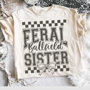 Op de afbeelding: Crèmekleurig T-shirt met de tekst "FERAL Ballfield SISTER" in verweerde grijze letters. Het ontwerp bevat een dambordpatroon en een strik. Het shirt is een casual, comfortabele stijl.