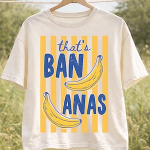 Puede incluir: Camiseta de color crema con un diseño divertido. La camiseta presenta la frase "that's BANANAS" en azul, con dos ilustraciones de plátanos amarillos. Rayas verticales amarillas sirven de fondo al texto y a los plátanos. La camiseta está colgada de una percha de madera.