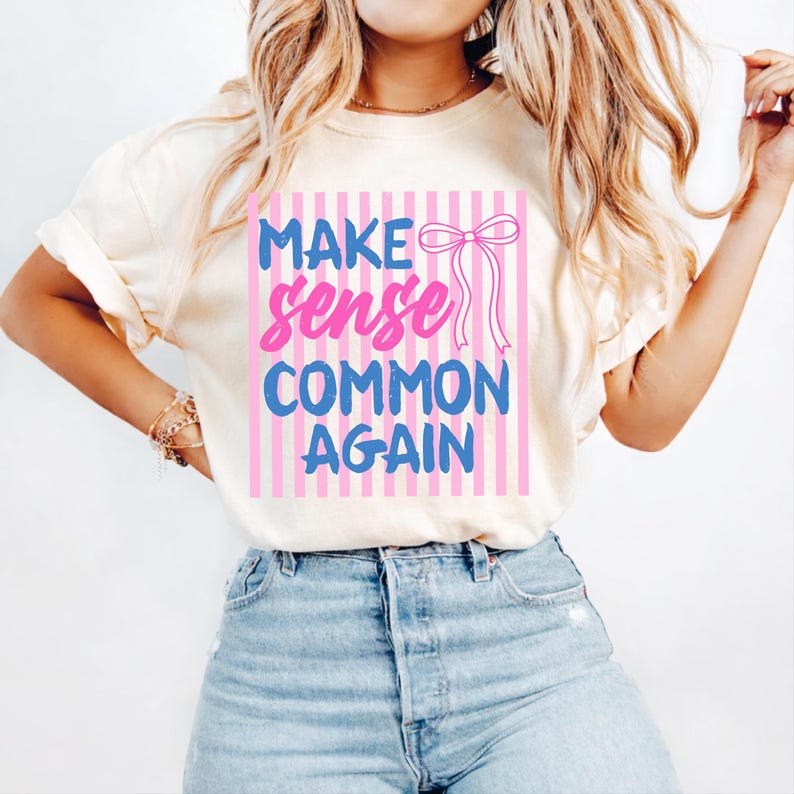 May include: Cream-colored t-shirt with the text "MAKE sense COMMON AGAIN" in blue and pink, with a bow graphic. The text is set against a background of pink and white vertical stripes. The shirt is paired with blue jeans.