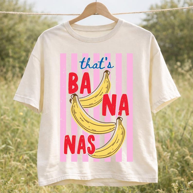 May include: Cream-colored t-shirt with a graphic design featuring three yellow bananas and the text "that's BANANAS" in red and blue. The design is set against a pink striped background.