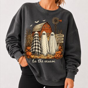 May include: A charcoal gray crewneck sweatshirt featuring a graphic of three ghosts standing in front of a red barn, a windmill, and corn stalks. The design includes orange pumpkins and a sign reading PUMPKINS, HAY-RIDES, CORN MAZE, with the text tis the season.