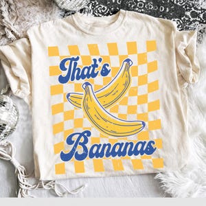 May include: Cream-colored t-shirt with a retro design featuring two yellow bananas and the text "That's Bananas" in blue script. The background has a yellow and white checkered pattern, giving it a vintage aesthetic.