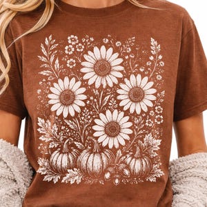 May include: A brown short-sleeved t-shirt featuring a white graphic print of three large sunflowers, smaller wildflowers, three pumpkins, and two acorns arranged in a rectangular composition on the front of the garment.