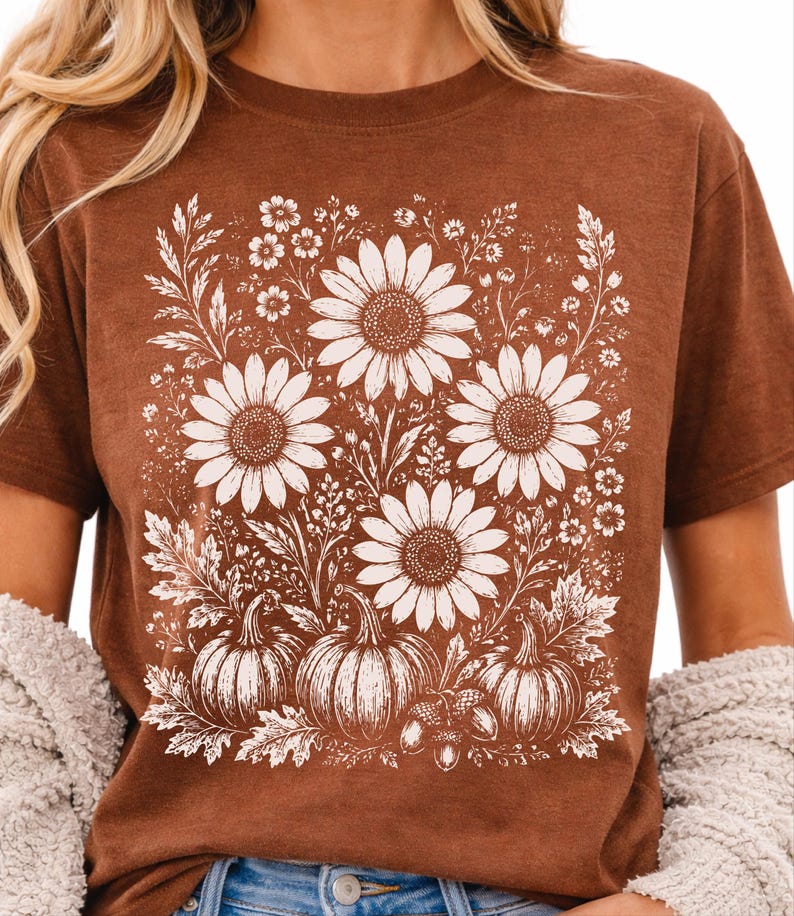 May include: A brown short-sleeved t-shirt featuring a white graphic print of three large sunflowers, smaller wildflowers, three pumpkins, and two acorns arranged in a rectangular composition on the front of the garment.